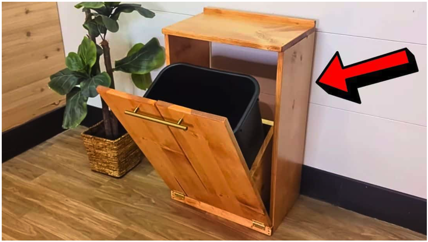 A wooden tilt-out trash bin cabinet with a black bin inside, cleverly designed to hide common eyesores, placed near a potted plant and against a wall with a red arrow pointing to it.