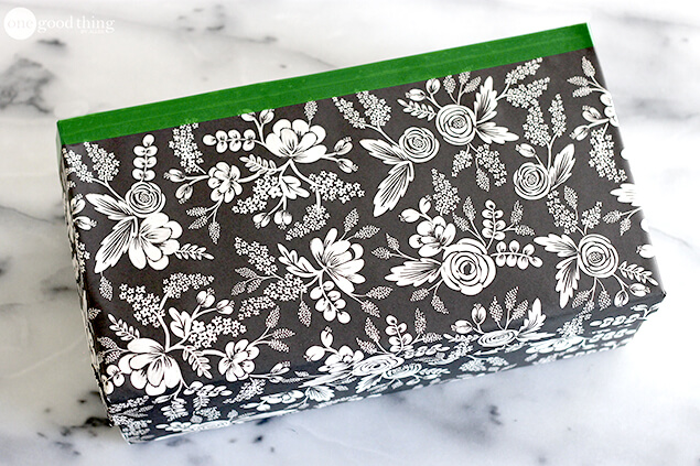 A rectangular box wrapped in black paper with a white floral pattern and green tape along the top edge sits on a marble surface, cleverly repurposed as a simple charging station for the whole family.