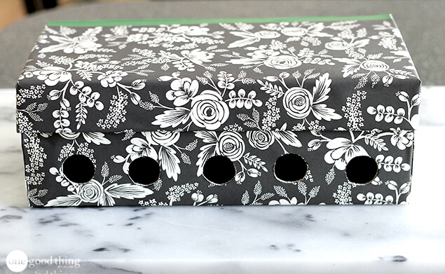 A decorative, black-and-white floral shoebox with six evenly spaced round holes cut in the side sits on a marble surface, repurposed as a simple charging station for organizing your family's devices.