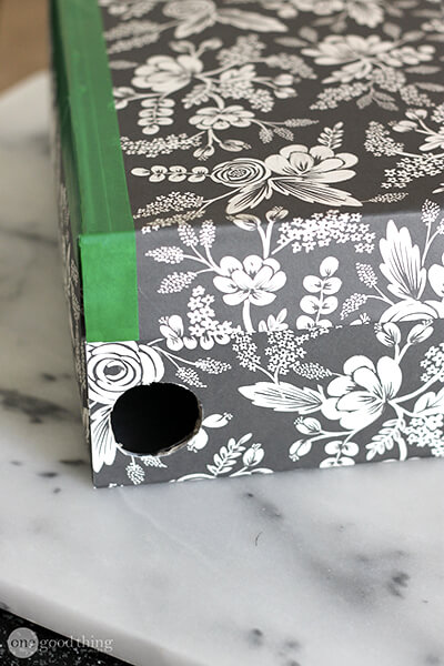 A black and white floral-covered box with a green edge and a circular hole on one side sits on a white marble surface, making for a stylish DIY charging station.