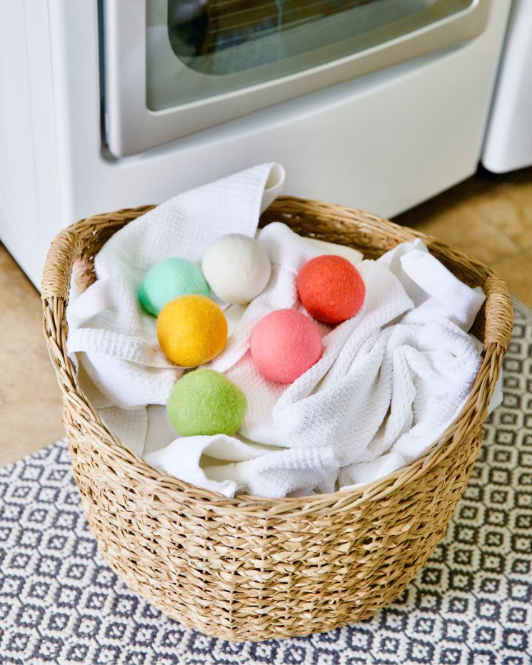 A wicker laundry basket filled with white towels and colorful wool dryer balls sits on a patterned rug in front of a washing machine, showing an easy way to use dryer balls for softer, fresher laundry.