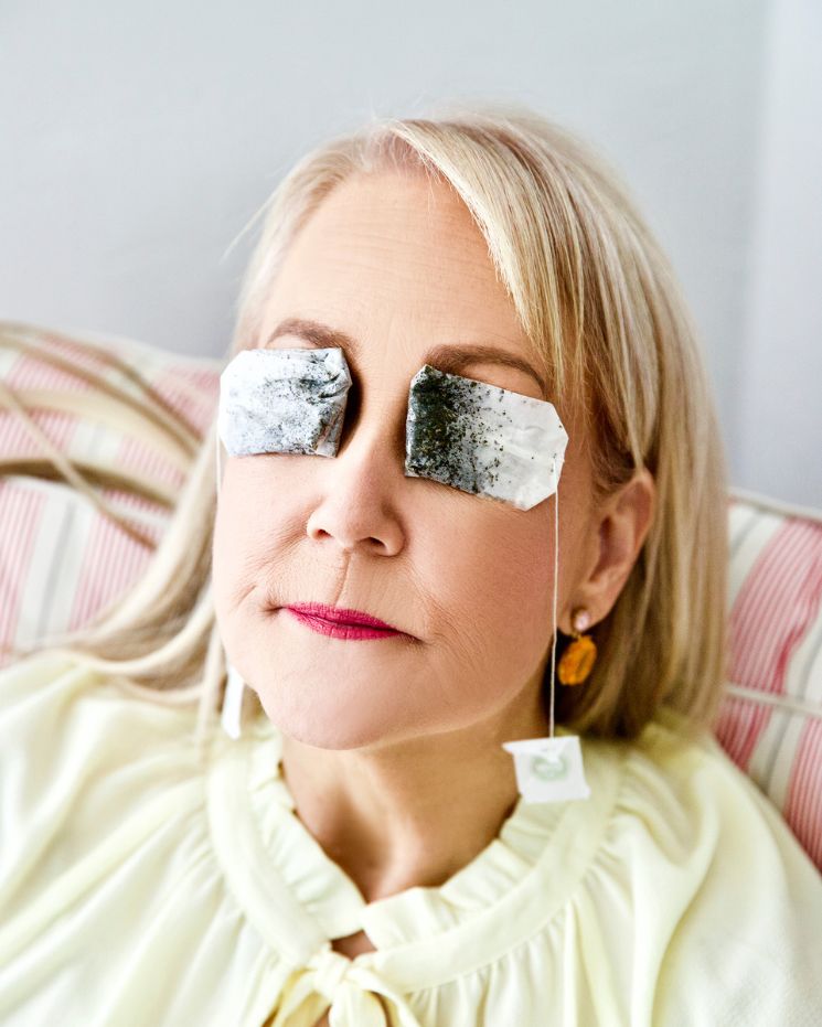 Woman with blonde hair sits on a striped couch with used tea bags over her closed eyes—a true beauty edition moment. She wears a light yellow top and gold earrings. Why didn’t I think of that?.