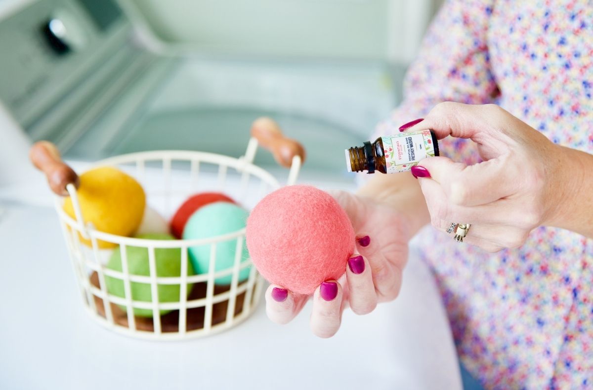 A person drips essential oil onto a pink wool dryer ball, showing how to use dryer balls with a basket of colorful ones and a washing machine in the background.