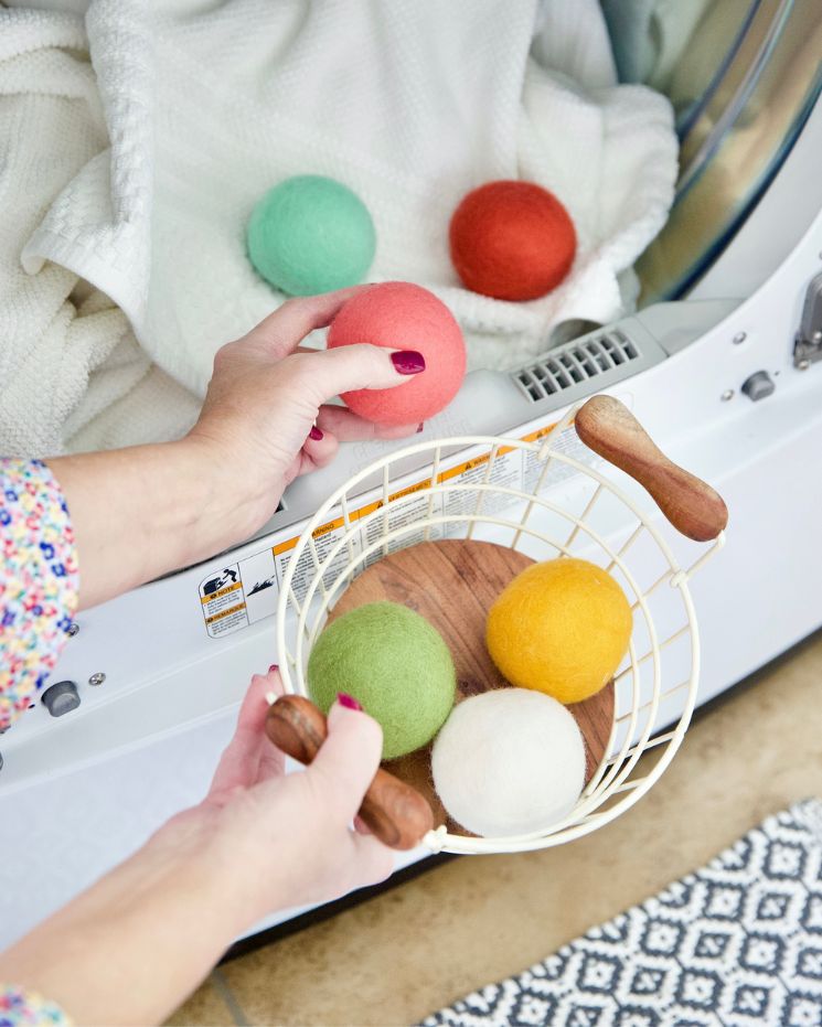 A person uses dryer balls, placing colorful wool ones into a dryer with a white blanket, while holding a basket filled with more vibrant dryer balls.