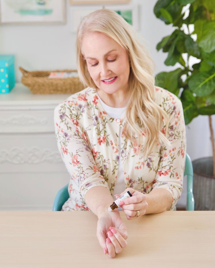 Woman applies essential oil to her wrist while seated at a table in a bright, decorated room with plants and home decor in the background, creating a soothing atmosphere that encourages a calm mind.