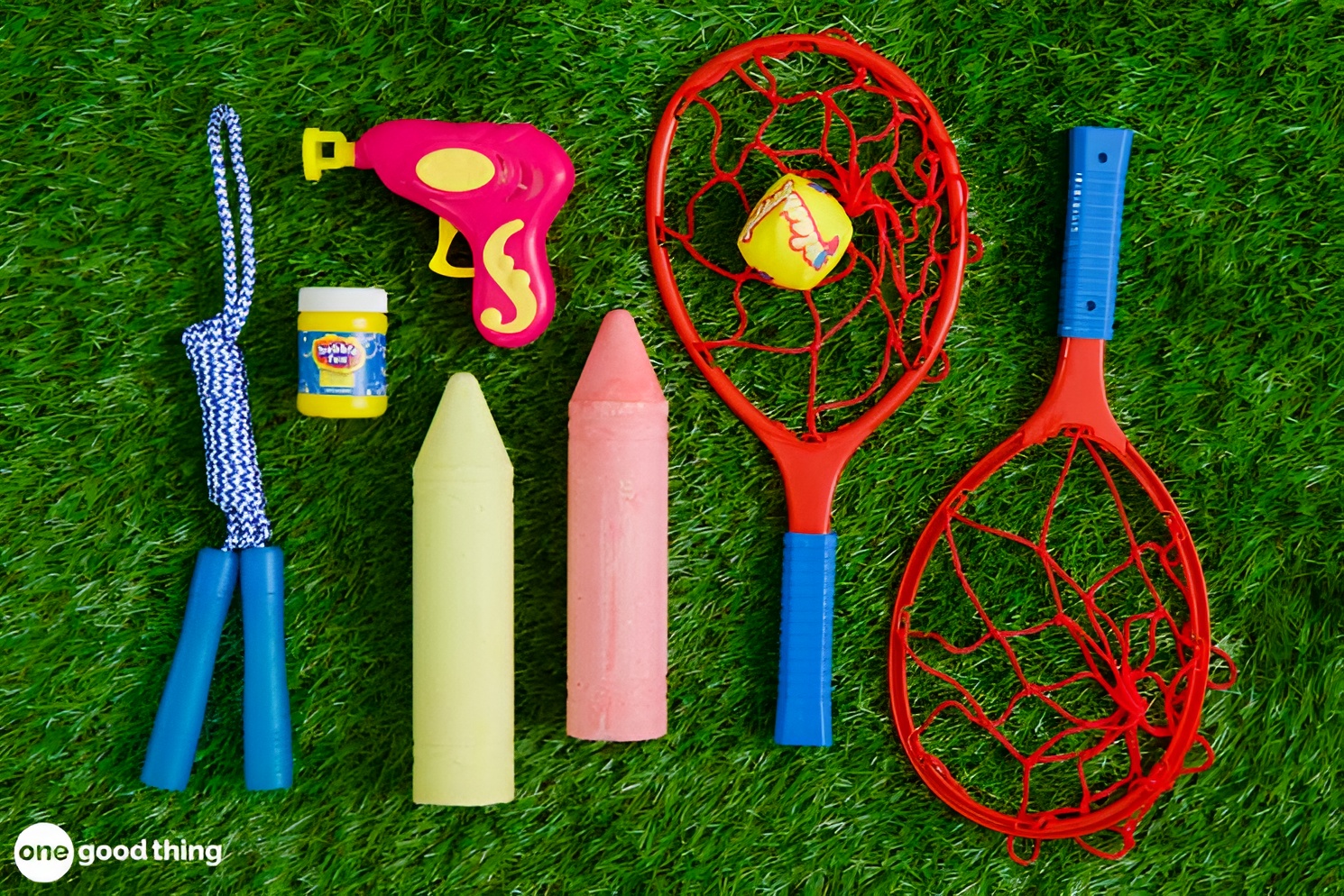 summer-dollar-store10 Children's outdoor toys from the dollar store, including a jump rope, bubble jar, yellow and pink chalk, a water gun, and two red plastic rackets with a yellow ball, are arranged on green grass.