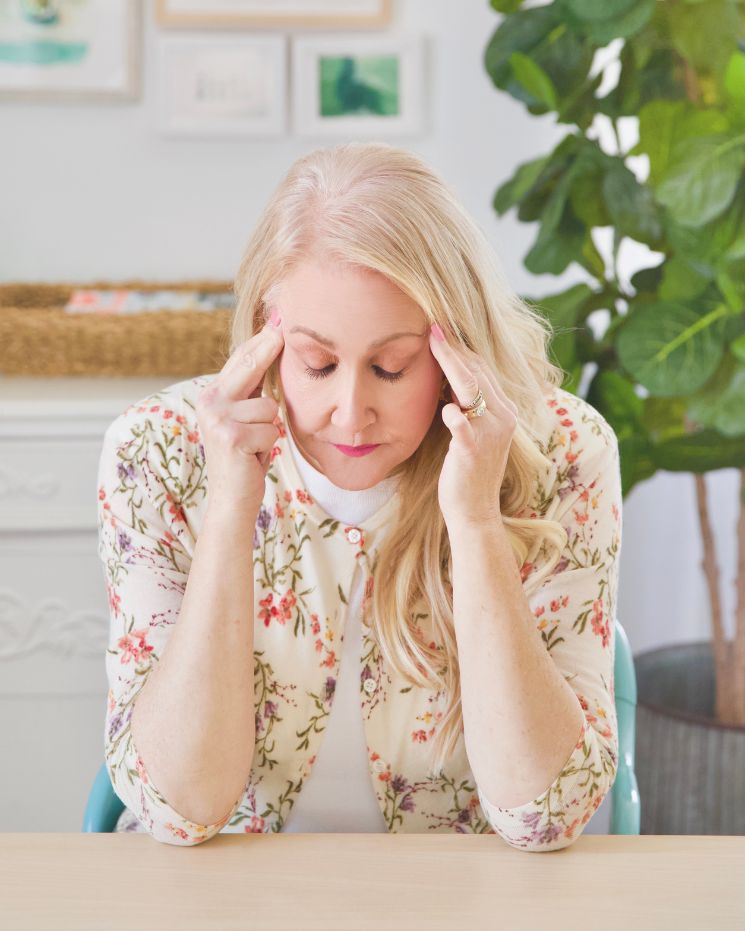 A woman with long blond hair sits at a table, touching her temples with both hands and looking down, appearing stressed or deep in thought as she seeks a calm mind.