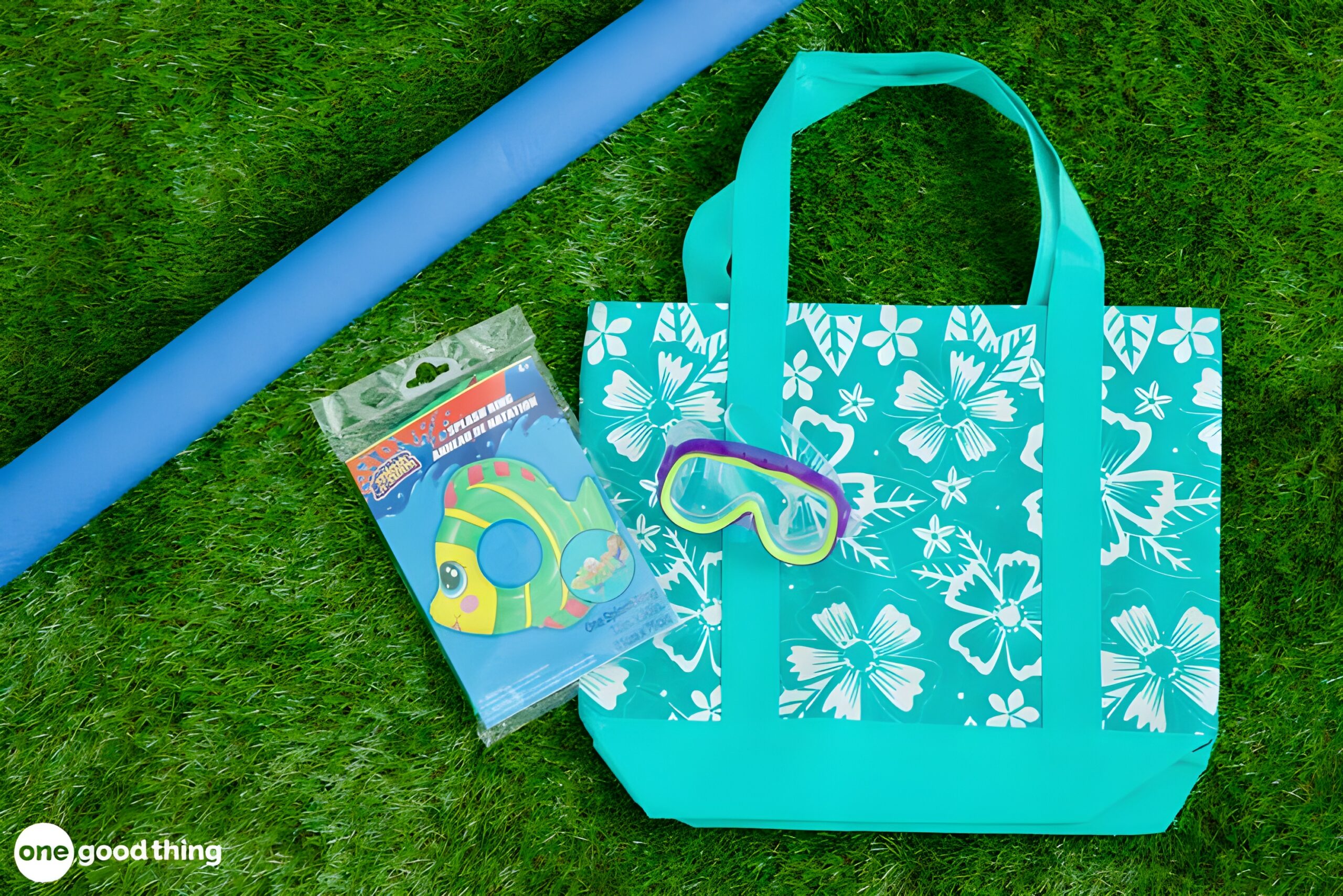 summer-dollar-store5 A blue floral tote bag, a blue pool noodle, kids' swim goggles, and a packaged inflatable fish float from the dollar store are arranged on green grass.