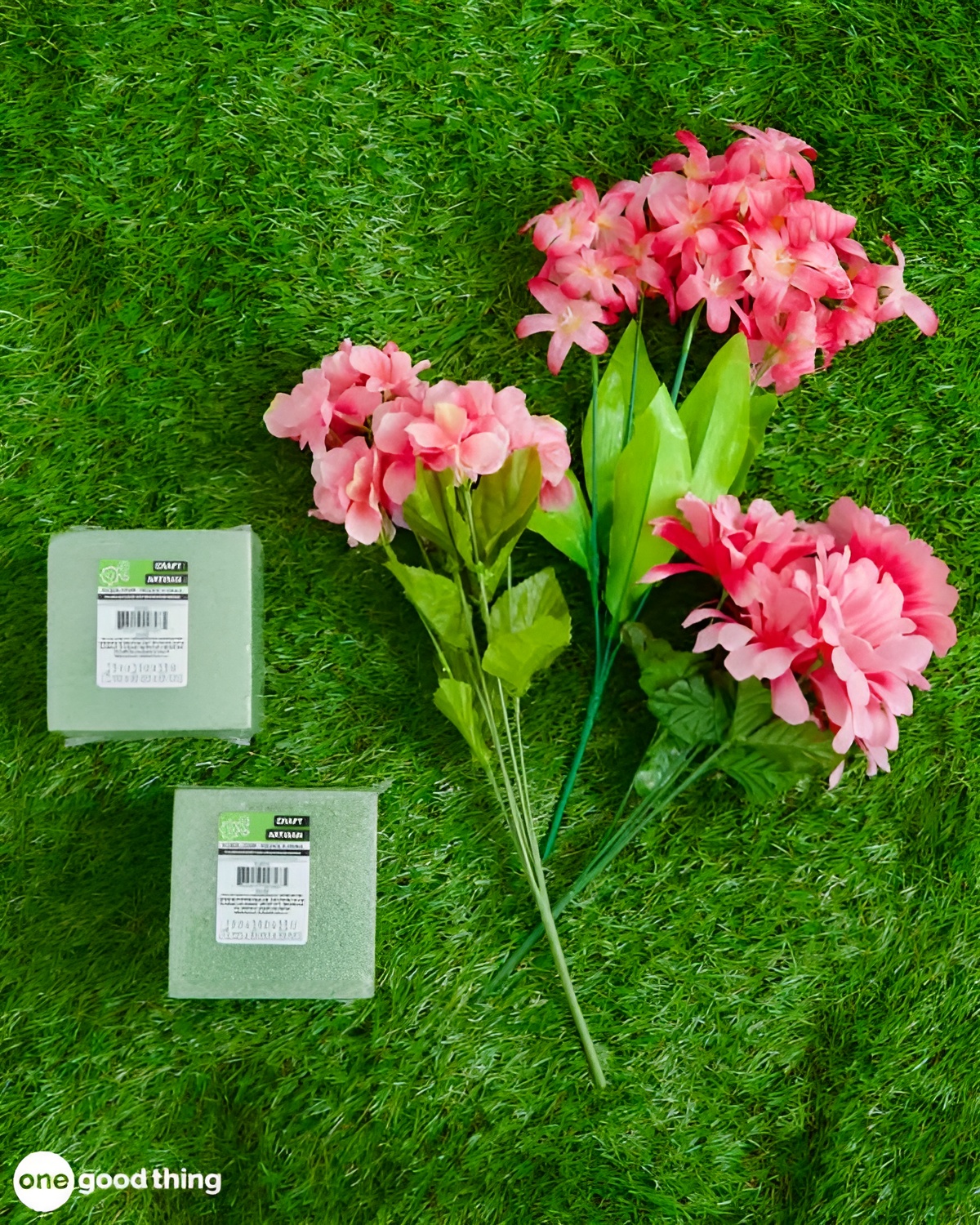 summer-dollar-store12-600x750 Two artificial pink flower stems and two green floral foam blocks, all sourced from the dollar store, are placed on a grass-like surface.