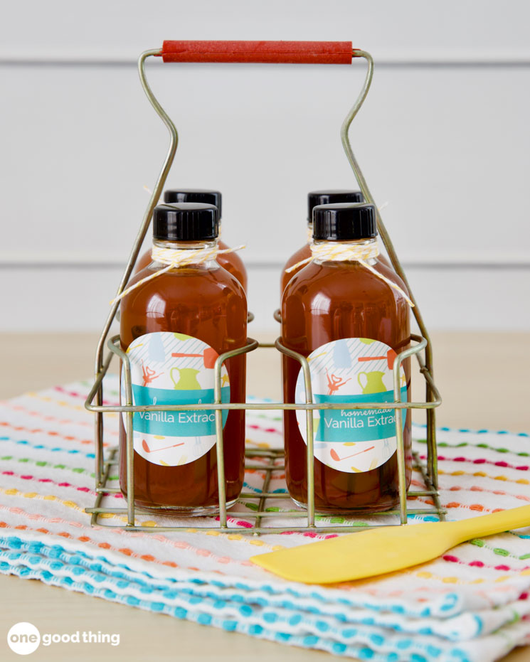 Four bottles labeled “Vanilla Extract” in a metal carrier sit on a colorful cloth with a yellow spatula beside them, making perfect Handmade Gifts for any occasion. This charming scene could easily be part of a Simple Gifts collection—perhaps even Part Five in your thoughtful series of homemade delights.