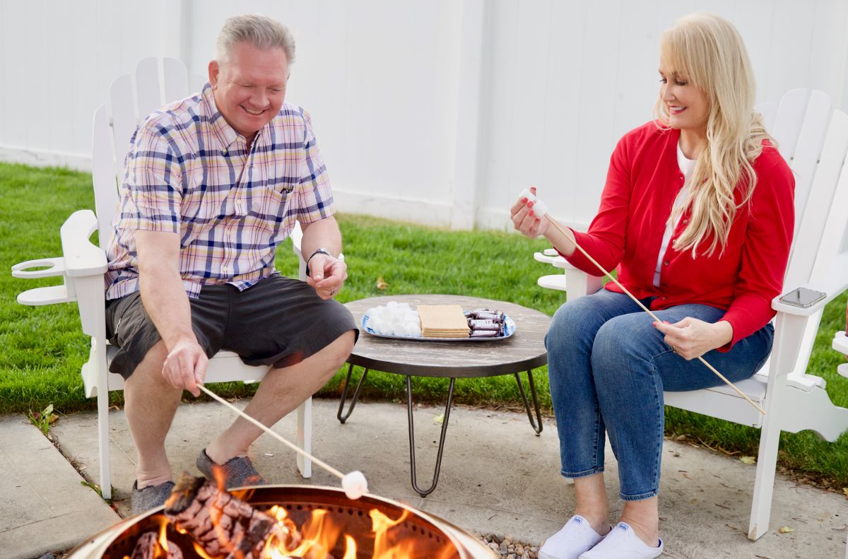soothe aches and pains_6 Two people sit in white chairs by a fire pit, gently easing their everyday aches as they roast marshmallows on sticks. A small table with graham crackers, chocolate, and marshmallows sits between them.