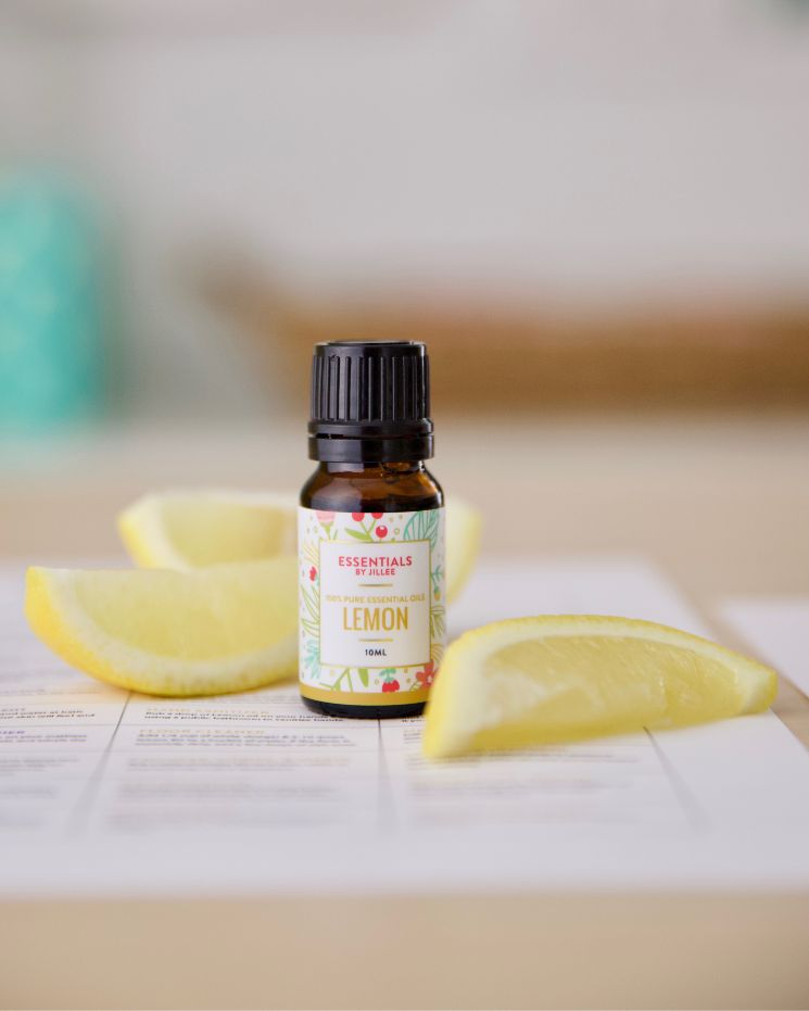 A small bottle labeled “Lemon Essential Oil” is placed on paper, surrounded by three lemon wedges on a light-colored surface—perfect for creating your own DIY goo gone solution.