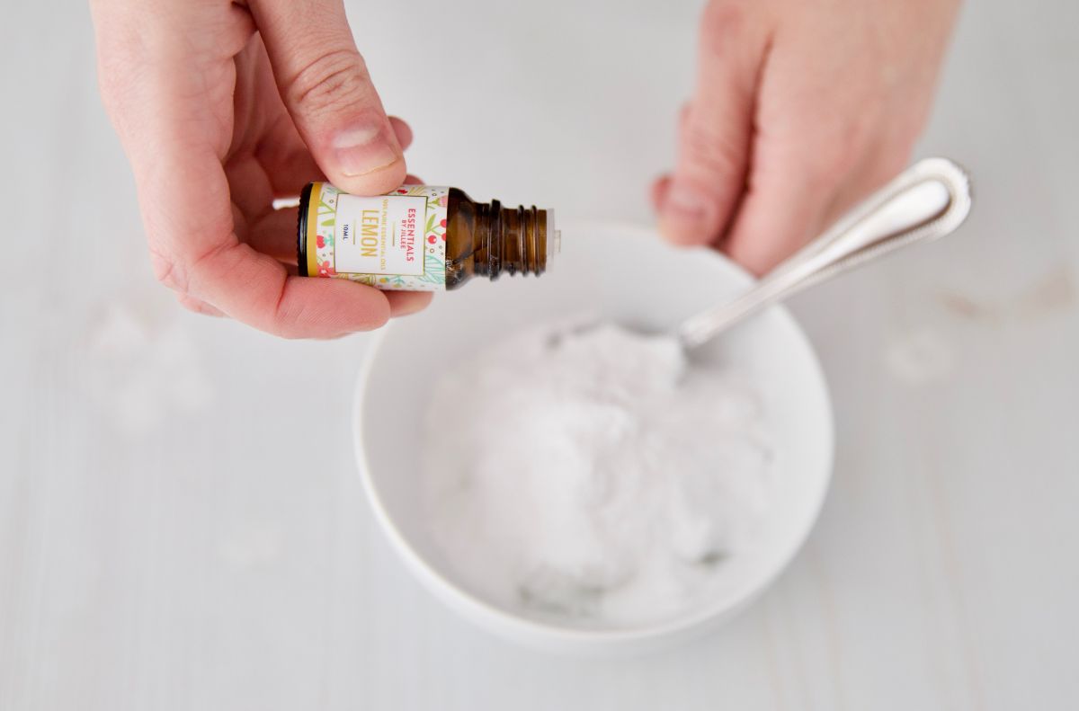 A person pours lemon essential oil from a small bottle into a white bowl filled with baking soda, holding a spoon in the bowl to create a homemade DIY goo gone solution.