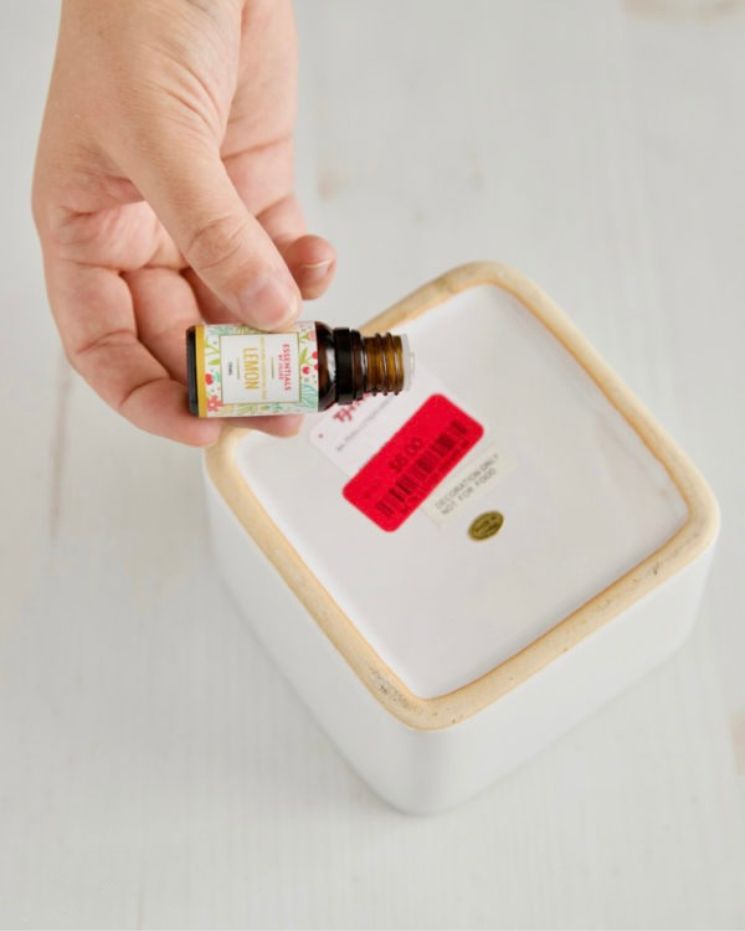 A hand holds a small essential oil bottle over a white ceramic container with a red price sticker on the bottom, perfect for creating your own DIY goo gone to easily remove sticky residue.